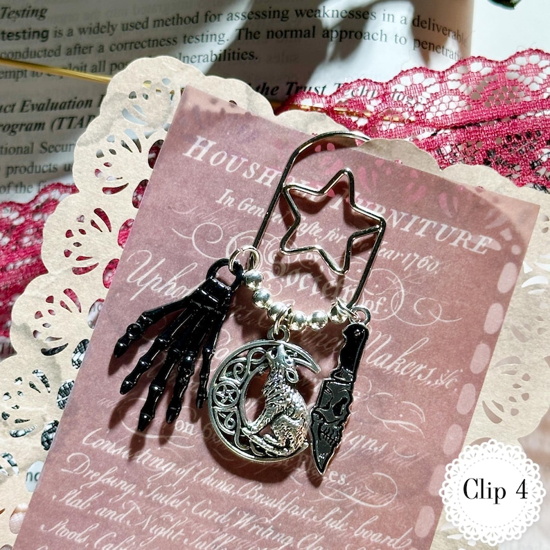 May include: A metal bookmark with a star-shaped top and decorative charms. Charms include a black skeleton hand, a crescent moon with a wolf, and a knife. The bookmark is on a pink paper with text.