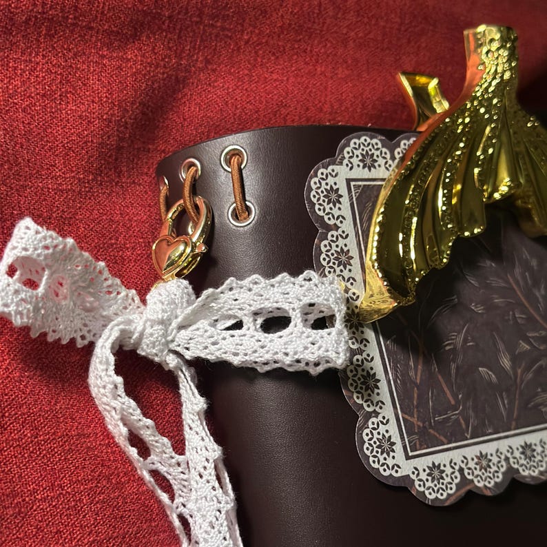 May include: A brown leather container with a white lace bow and gold-colored accents. The container has a decorative gold charm and a patterned label. The background is a solid red color.