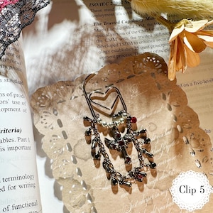 May include: A silver-toned metal bookmark with a heart-shaped top and dangling charms. The charms include small beads, crosses, and red gemstones. The bookmark is resting on a decorative paper doily and a book. The text "Clip 5" is visible.