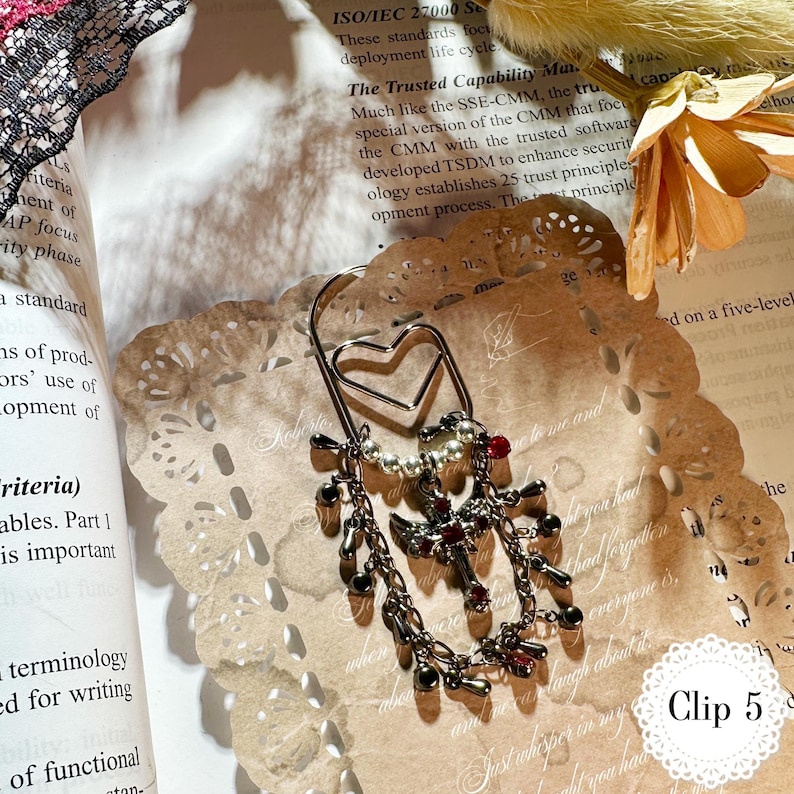 May include: A silver-toned metal bookmark with a heart-shaped top and dangling charms. The charms include small beads, crosses, and red gemstones. The bookmark is resting on a decorative paper doily and a book. The text "Clip 5" is visible.
