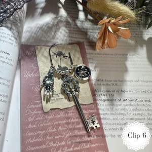May include: A silver bookmark with three charms: a hand, a key, and a circular emblem. The key charm is detailed with a fleur-de-lis design. The bookmark is displayed on a book page with text and a pink card.