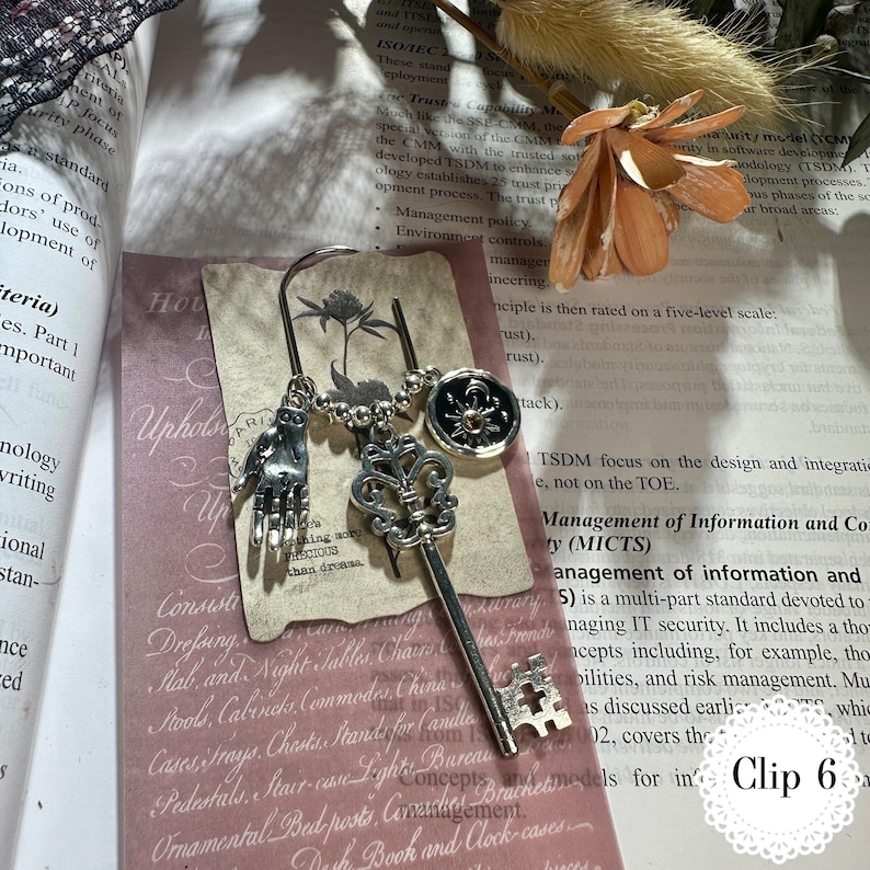 May include: A silver bookmark with three charms: a hand, a key, and a circular emblem. The key charm is detailed with a fleur-de-lis design. The bookmark is displayed on a book page with text and a pink card.