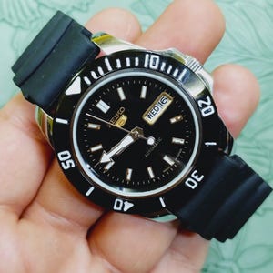Pode incluir: Um relógio automático Seiko preto com mostrador preto, algarismos brancos e pulseira de borracha preta. O mostrador mostra o dia e a data. A moldura tem algarismos brancos em intervalos de 5 minutos. O relógio é segurado em uma mão.
