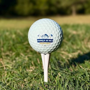 Puede incluir: Una pelota de golf blanca con una superficie con hoyuelos, colocada sobre un tee blanco, sobre un fondo de hierba verde y un fondo borroso. La pelota de golf tiene el texto "SHANKS R US GOLF" en azul. El tee está en el centro del encuadre.