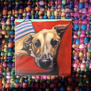 May include: A painting of a brown and tan dog with a pink and white striped pillow behind it. The dog is lying on a red couch and looking directly at the viewer.