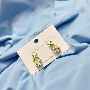 May include: Gold-toned earrings with a teardrop design, featuring a large, clear crystal at the center and smaller crystals along the curved frame. The earrings are displayed on a white card with the text "Fashion jewelry" and "MADE IN CHINA".