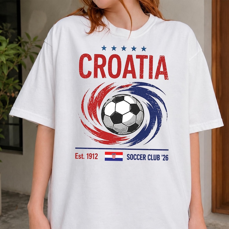 K&ouml;nnte beinhalten: Ein wei&szlig;es T-Shirt mit dem roten Schriftzug CROATIA &uuml;ber einer Grafik eines Fu&szlig;balls, der von roten und blauen Wirbellinien umgeben ist. Unter dem Ball stehen der Text Est. 1912, eine kleine kroatische Flagge und SOCCER CLUB '26 mit f&uuml;nf blauen Sternen.