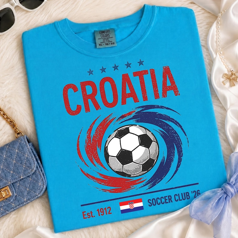 K&ouml;nnte beinhalten: Ein hellblaues T-Shirt mit dem Wort CROATIA in roten Buchstaben &uuml;ber einer Grafik eines Fu&szlig;balls, der von roten und blauen Wirbeln umgeben ist. Unter dem Ball stehen der Text Est. 1912, eine kleine kroatische Flagge und SOCCER CLUB '26.