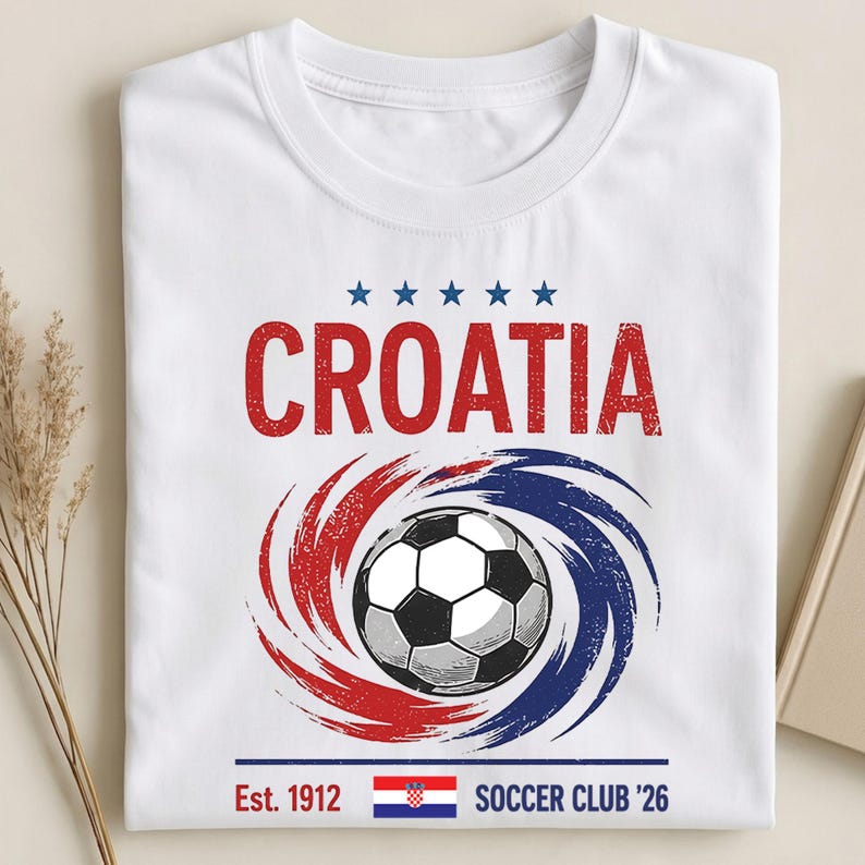 K&ouml;nnte beinhalten: Ein wei&szlig;es T-Shirt mit einem Aufdruck, der das Wort CROATIA in Rot, f&uuml;nf blaue Sterne, einen Fu&szlig;ball in einem rot-blauen Wirbel sowie den Text Est. 1912 mit der kroatischen Flagge und den Worten SOCCER CLUB '26 am unteren Rand zeigt.