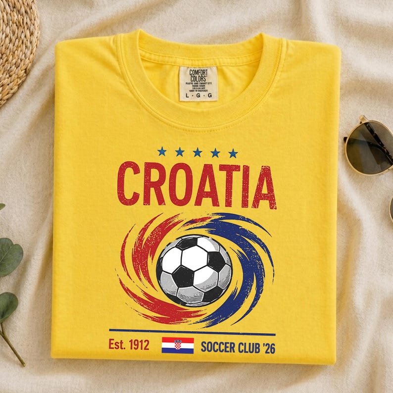 K&ouml;nnte beinhalten: Ein gelbes Kurzarm-T-Shirt mit dem roten Schriftzug CROATIA, einem zentralen Fu&szlig;ball-Motiv umgeben von roten und blauen Wirbeln, f&uuml;nf blauen Sternen sowie dem Text Est. 1912, einer kleinen kroatischen Flagge und SOCCER CLUB '26 unten.
