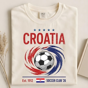 K&ouml;nnte beinhalten: Ein cremefarbenes T-Shirt mit einer Grafik, die das Wort CROATIA in Rot, f&uuml;nf blaue Sterne, einen Fu&szlig;ball &uuml;ber roten und blauen wirbelnden Pinselstrichen, den Text Est. 1912, die kroatische Flagge und den Text SOCCER CLUB '26 zeigt.