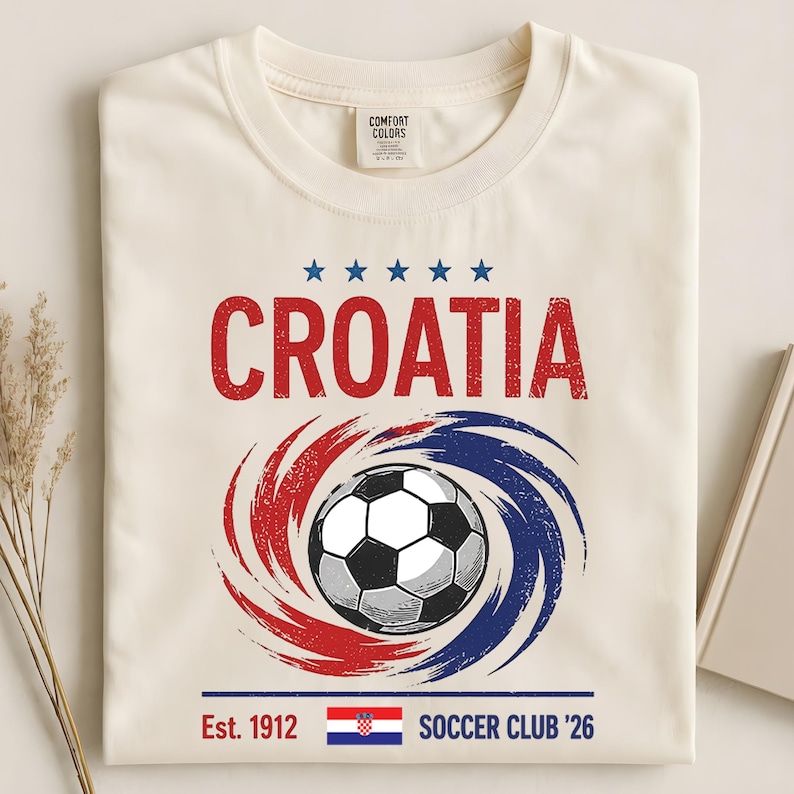 K&ouml;nnte beinhalten: Ein cremefarbenes T-Shirt mit einer Grafik, die das Wort CROATIA in Rot, f&uuml;nf blaue Sterne, einen Fu&szlig;ball &uuml;ber roten und blauen wirbelnden Pinselstrichen, den Text Est. 1912, die kroatische Flagge und den Text SOCCER CLUB '26 zeigt.