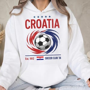 K&ouml;nnte beinhalten: Ein wei&szlig;er Kapuzenpullover mit einer Grafik, die den roten Schriftzug CROATIA, f&uuml;nf blaue Sterne, einen schwarz-wei&szlig;en Fu&szlig;ball umgeben von roten und blauen Wirbeln sowie den Text Est. 1912, eine kroatische Flagge und SOCCER CLUB '26 zeigt.