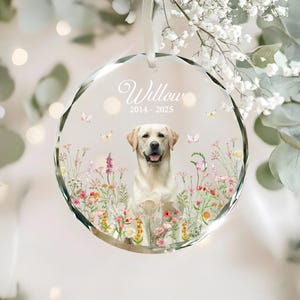 May include: A clear, faceted glass ornament with a Labrador Retriever portrait, surrounded by colorful wildflowers and butterflies. The ornament has the name "Willow" and dates "2014-2025" in elegant script, suspended by a white ribbon.
