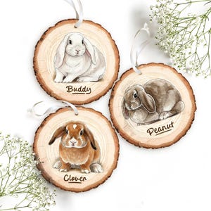 Puede incluir: Tres adornos de madera redondos con ilustraciones de acuarela de conejos. Cada adorno presenta un conejo diferente: un conejo blanco llamado "Buddy", un conejo marrón llamado "Peanut" y un conejo marrón y blanco llamado "Clover". Cada adorno tiene una cinta blanca para colgar.
