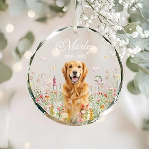 May include: A clear, faceted glass ornament with a golden retriever illustration. The ornament features the name "Marley" and the years "2016-2025". The dog is surrounded by colorful flowers and butterflies, with a white ribbon for hanging.