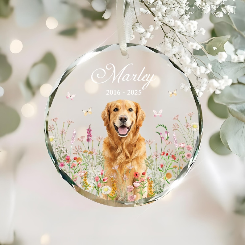 May include: A clear, faceted glass ornament with a golden retriever illustration. The ornament features the name "Marley" and the years "2016-2025". The dog is surrounded by colorful flowers and butterflies, with a white ribbon for hanging.