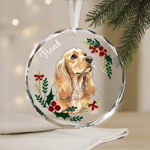 May include: A clear, round Christmas ornament with a Cocker Spaniel illustration. The dog is light brown with long, wavy ears. The ornament has the name "Hank" in script, with holly and red bow accents.