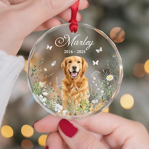 May include: A clear, round ornament with a red ribbon. The ornament features a golden retriever illustration surrounded by flowers and butterflies. The name "Marley" and the years "2016-2025" are displayed on the ornament.