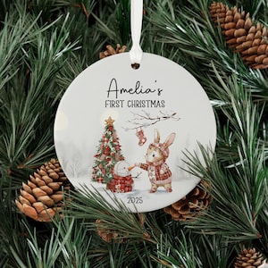 Może przedstawiać: Okrągła, biała ozdoba świąteczna z napisem "Amelia's FIRST CHRISTMAS" i rokiem 2025. Ozdoba przedstawia świąteczną ilustrację królika, bałwana i choinki. Do zawieszenia przymocowana jest biała wstążka.