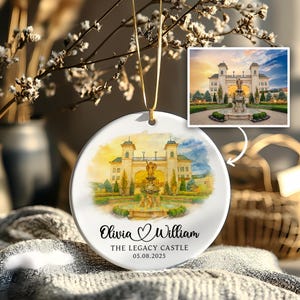 May include: A white ceramic ornament with a watercolor painting of a castle and fountain. The ornament has a gold string for hanging and features the names "Olivia & William" and the date "05.08.2025".
