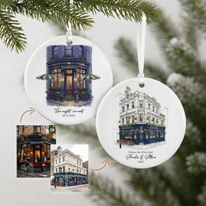 May include: Two white ceramic Christmas ornaments with illustrations of buildings. One features a blue building with green and white striped awnings, the text "The night we met" and a date. The other shows a white building with blue accents and the text "Where We First Met".