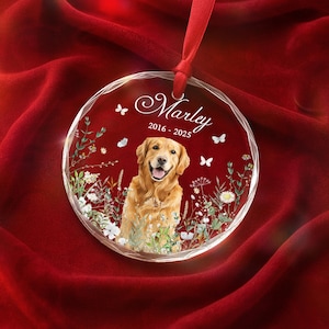 May include: A clear, round ornament with a smiling golden retriever image, surrounded by floral accents and butterflies. The ornament has the name "Marley" and dates "2016-2025" in elegant script, suspended by a red ribbon.