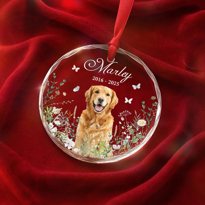 May include: A clear, round ornament with a smiling golden retriever image, surrounded by floral accents and butterflies. The ornament has the name "Marley" and dates "2016-2025" in elegant script, suspended by a red ribbon.