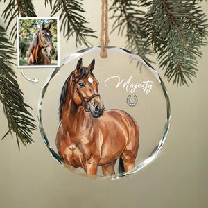 May include: A round, clear glass ornament with a watercolor painting of a brown horse wearing a bridle. The word "Majesty" is written in cursive above a horseshoe. A smaller image of the horse is in the upper left corner.