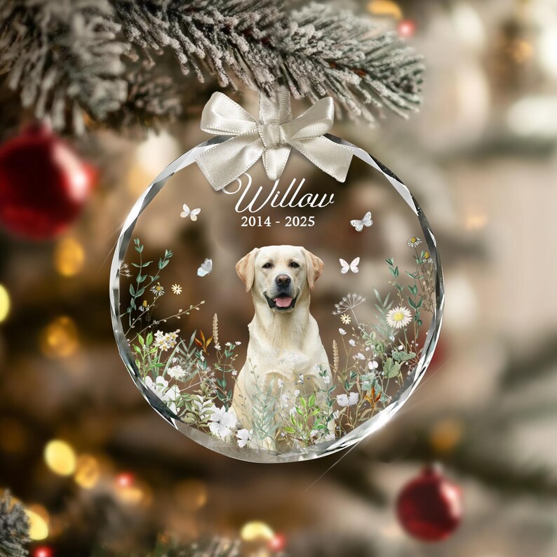 May include: A clear, faceted glass Christmas ornament with a cream-colored Labrador Retriever inside. The ornament has the name "Willow" and the years "2014-2025" printed on it. White butterflies and flowers decorate the inside, and a white bow is at the top.