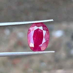 May include: A faceted, oval-shaped ruby gemstone held by silver tweezers. The ruby is a deep red color, with visible internal facets reflecting light. The background is blurred, suggesting an outdoor setting.