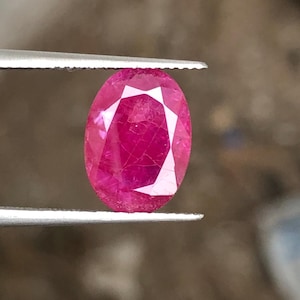 May include: A faceted, oval-shaped ruby gemstone held by tweezers. The ruby is a vibrant shade of red with visible internal structures and facets that catch the light. The tweezers are silver-colored.