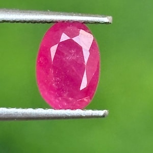 May include: A vibrant, oval-cut pinkish-red gemstone held securely between the two metal tips of a pair of silver-toned tweezers. The faceted stone displays a brilliant, polished surface with clear geometric cuts reflecting light.