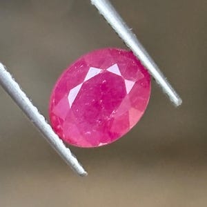 May include: A vibrant, oval-cut ruby gemstone with a faceted surface that reflects light. The deep pinkish-red stone is held securely by a pair of silver-toned metal tweezers, showcasing its translucent quality and precise geometric cut.