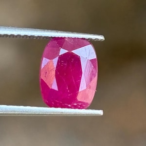 以下が含まれることがあります： 鮮やかなクッションカットのルビーで、深い赤色と多面的な表面を持ち、ピンセットの2本の細い金属製の先端の間にしっかりと保持され、その半透明の透明度と磨かれた幾何学的なエッジを示しています。
