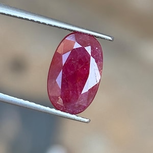 May include: A faceted oval-shaped ruby gemstone with a deep red color held by a pair of silver-toned metal tweezers. The gemstone displays multiple triangular and kite-shaped facets across its surface, reflecting light with a clear, polished finish.