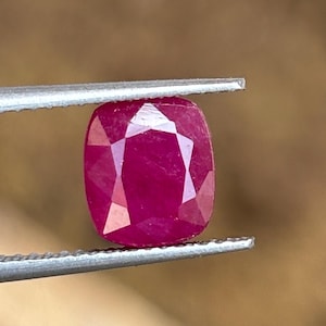 May include: A cushion-cut ruby gemstone with a deep red colour and faceted surface held by a pair of metal tweezers. The gemstone displays a clear geometric cut with multiple triangular and trapezoidal facets reflecting light across its surface.