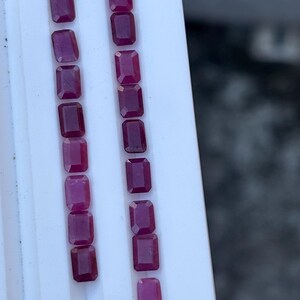 Op de afbeelding: Twee parallelle rijen rechthoekige, dieprode edelstenen worden weergegeven op een wit oppervlak. De edelstenen hebben een gepolijst, reflecterend oppervlak en zijn gelijkmatig verdeeld. De belichting benadrukt de kleur en helderheid van de stenen.