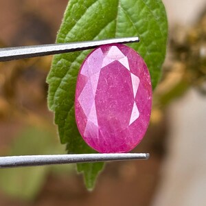 Könnte beinhalten: Ein facettierter, ovaler Rubin-Edelstein, der von einer Pinzette gehalten wird. Der Rubin hat eine leuchtende, rötlich-rosa Farbe mit mehreren Facetten, die das Licht reflektieren. Der Hintergrund umfasst ein grünes Blatt und verschwommenes Laub.