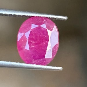 May include: A vibrant, oval-cut, deep pink gemstone held by a pair of metal tweezers. The stone features a faceted surface that reflects light, showcasing a saturated, translucent color throughout its polished, symmetrical oval shape.