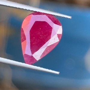 May include: A faceted, pear-shaped ruby gemstone held by tweezers. The ruby displays a deep red colour with multiple facets catching the light. The tweezers are silver and the background is blurred.