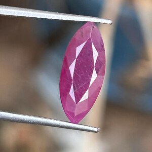 May include: A marquise-cut ruby gemstone held by tweezers. The ruby is a deep pinkish-red color with facets that reflect light. The gemstone is elongated and has a pointed end on each side.