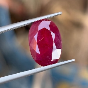 May include: A vibrant, oval-cut ruby gemstone held by silver tweezers. The ruby displays a deep red hue and multiple facets, reflecting light. The gemstone is the central focus, highlighting its clarity and brilliance.