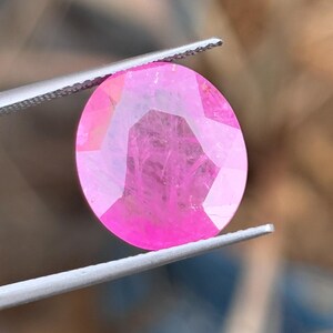 Puede incluir: Una gema rosa ovalada y facetada es sostenida por unas pinzas. La gema tiene un corte brillante, reflejando la luz y mostrando su estructura interna. El fondo está borroso, enfatizando el color y la claridad de la gema.