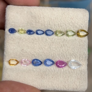 May include: A collection of pear-shaped gemstones in various colors, including blue, yellow, pink, and clear. The gems are arranged in two rows on a beige fabric, showcasing their different hues and clarity. The gemstones are likely for jewelry making.