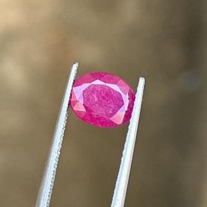 May include: A vibrant, oval-cut ruby gemstone with a faceted surface, displaying a deep pinkish-red hue. The gemstone is held securely between the two metal tips of a pair of silver-toned tweezers, showcasing its clarity and color.
