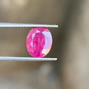 May include: A vibrant, oval-cut ruby gemstone with a deep pinkish-red hue and multifaceted surface, held securely between the two metal prongs of a pair of tweezers. The stone displays internal clarity and light reflection across its facets.