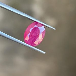 May include: A vibrant, oval-cut ruby gemstone with a faceted surface that reflects light, held securely by a pair of silver-toned metal tweezers. The gemstone displays a deep, saturated pinkish-red hue and is positioned centrally in the frame.