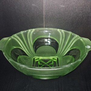 May include: A green glass bowl with a scalloped rim and two handles. The bowl has a geometric design on the base and a fan-like pattern radiating from the centre. The bowl is made of a translucent green glass.