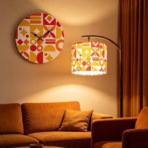 May include: A round wall clock and a floor lamp with a matching drum shade, both featuring a geometric pattern in shades of red, orange, and yellow. The clock has black hands, and the lamp has a curved metal arm emitting a warm light.